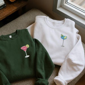 May include: Two sweatshirts, one dark green and one white, are displayed. Each has a cocktail graphic on the front. The green sweatshirt features a pink margarita with a lime. The white sweatshirt has a blue and yellow cocktail with a sprig of greenery.