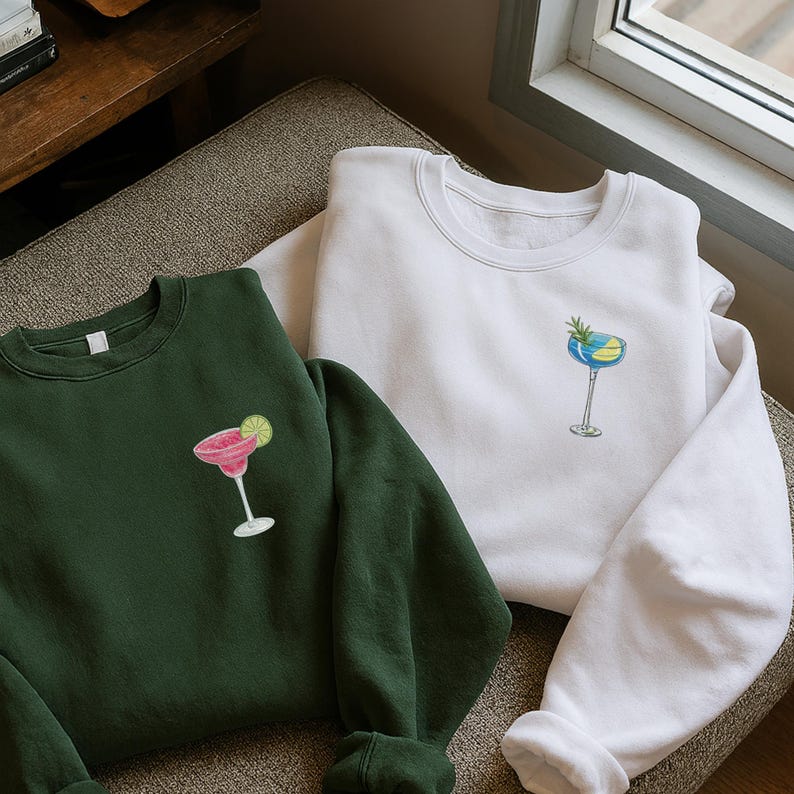 May include: Two sweatshirts, one dark green and one white, are displayed. Each has a cocktail graphic on the front. The green sweatshirt features a pink margarita with a lime. The white sweatshirt has a blue and yellow cocktail with a sprig of greenery.