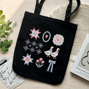 May include: Black tote bag with embroidered designs including stars, flowers, and two geese with bows. The bag has two handles and is made of a sturdy fabric. The design is in pastel colors.
