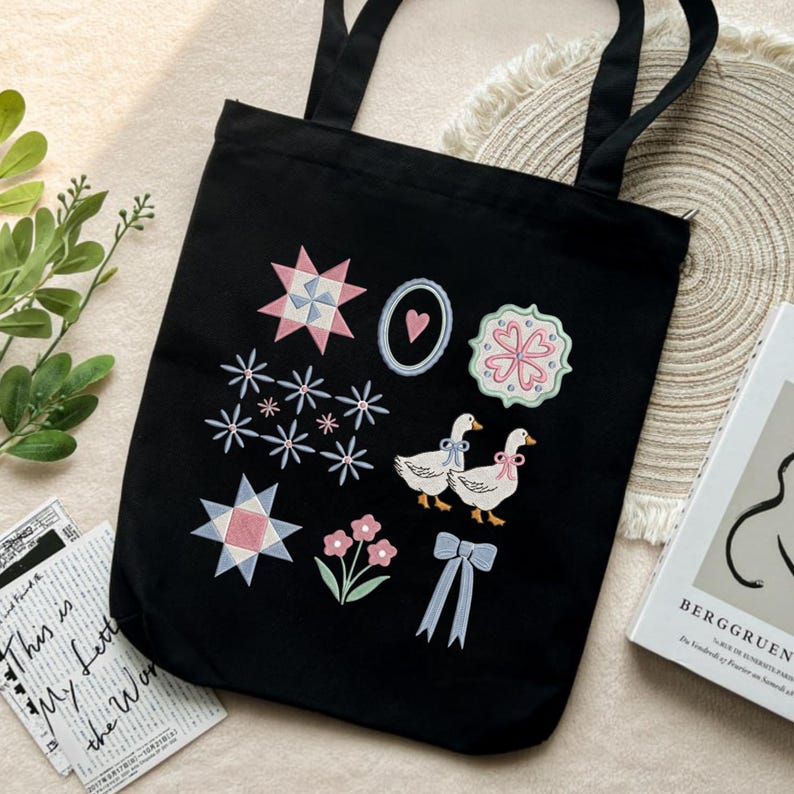 May include: Black tote bag with embroidered designs including stars, flowers, and two geese with bows. The bag has two handles and is made of a sturdy fabric. The design is in pastel colors.