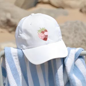 May include: A white baseball cap with a strawberry patch design. The strawberry is made of various patterned fabrics in shades of pink, red, and green. The cap is resting on a blue and white striped towel.