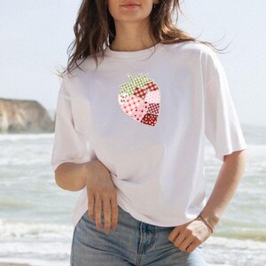 May include: White t-shirt featuring a patchwork strawberry design. The strawberry is composed of various patterned fabrics in shades of red, pink, and green. The shirt has short sleeves and a relaxed fit, suitable for casual wear.
