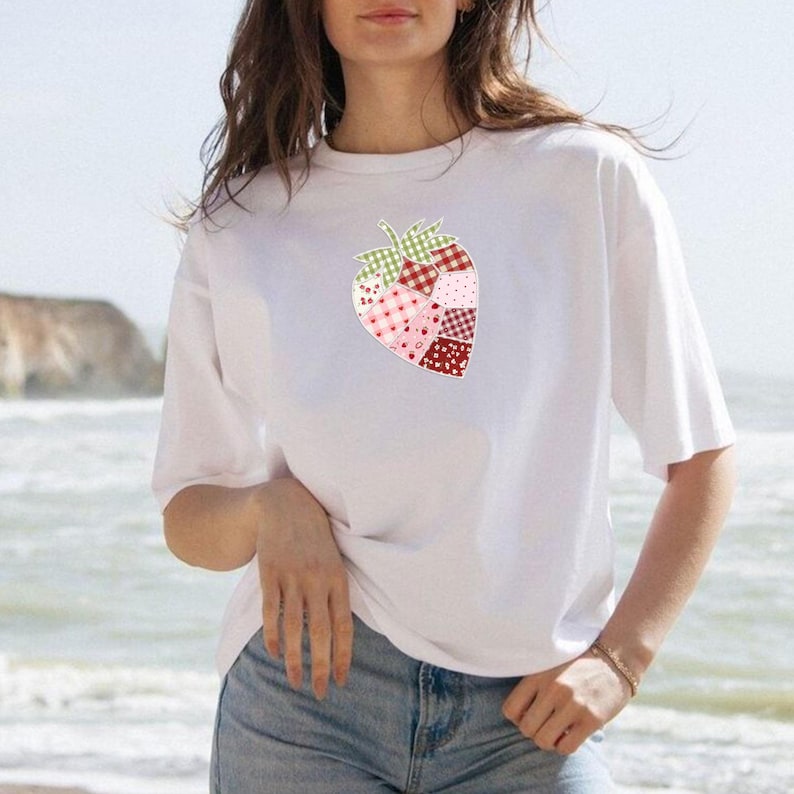 May include: White t-shirt featuring a patchwork strawberry design. The strawberry is composed of various patterned fabrics in shades of red, pink, and green. The shirt has short sleeves and a relaxed fit, suitable for casual wear.