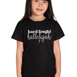 K&ouml;nnte beinhalten: Schwarzes T-Shirt mit dem wei&szlig;en Schriftzug "hard fought hallelujah". Das T-Shirt wird von einer jungen Person getragen. Der Text enth&auml;lt ein Kreuzsymbol.