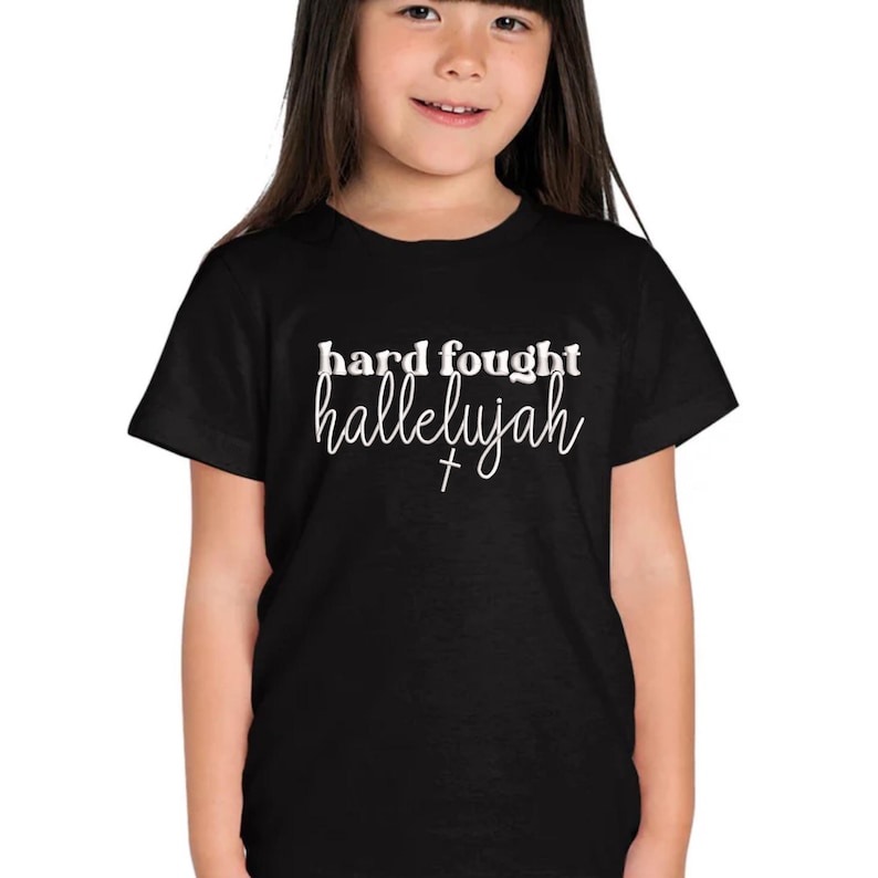 K&ouml;nnte beinhalten: Schwarzes T-Shirt mit dem wei&szlig;en Schriftzug "hard fought hallelujah". Das T-Shirt wird von einer jungen Person getragen. Der Text enth&auml;lt ein Kreuzsymbol.