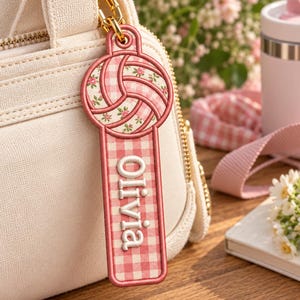 Könnte beinhalten: Ein rechteckiger bestickter Schlüsselanhänger mit einer Volleyball-Grafik an der Oberseite in einem rosa-weißen Karomuster mit kleinen floralen Akzenten. Der untere Teil hat einen rosa-weißen Karohintergrund mit dem Namen Olivia in Weiß.
