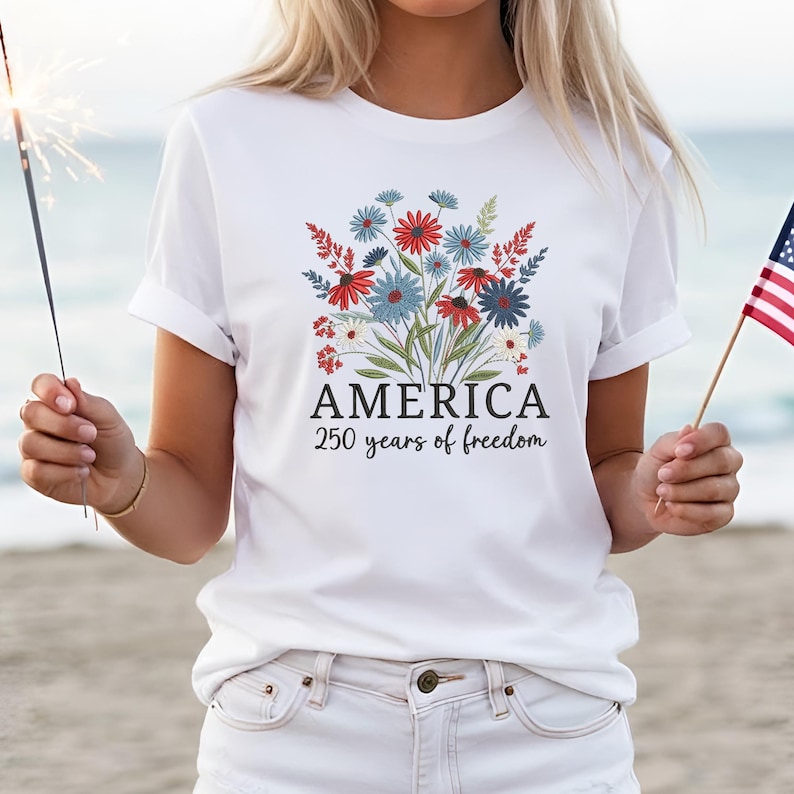 Puede incluir: Camiseta blanca con un dise&ntilde;o floral en rojo, blanco y azul. La camiseta presenta la palabra "AMERICA" y el texto "250 years of freedom" en negro. La camiseta es un dise&ntilde;o patri&oacute;tico.