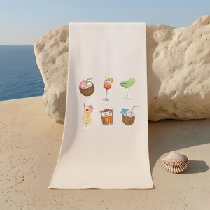 May include: A cream-colored beach towel featuring watercolor illustrations of six tropical cocktails. The drinks include a coconut with a flower, a cocktail in a stemmed glass with an orange slice, and a margarita with a lime wedge.