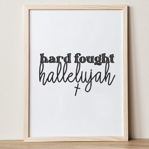 K&ouml;nnte beinhalten: Gerahmter Druck mit den Worten "hard fought hallelujah" in schwarzer Schrift und einem kleinen Kreuz. Die Worte sind auf wei&szlig;em Hintergrund zentriert, der Rahmen ist helles Holz.
