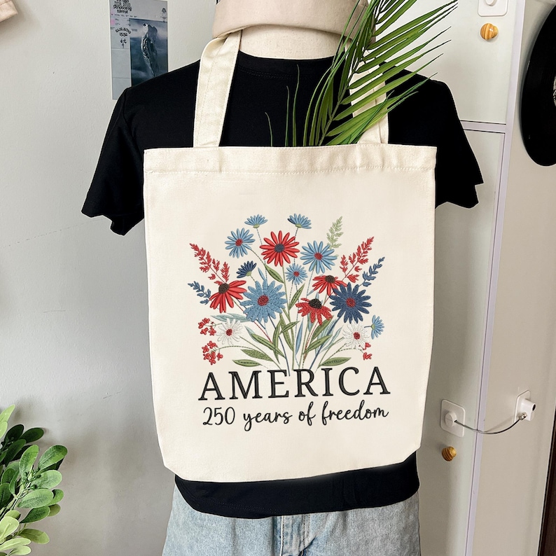 Puede incluir: Bolso tote de color crema con un dise&ntilde;o floral en rojo, blanco y azul. La palabra "AMERICA" est&aacute; impresa debajo de las flores, con "250 years of freedom" debajo. El bolso tiene dos asas de color crema.