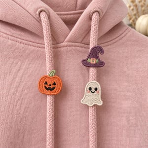 Op de afbeelding: Een oudroze hoodie met twee bijpassende trekkoorden. Aan het ene trekkoord zit een geborduurde oranje pompoenpatch, terwijl het andere een paarse heksenhoedpatch heeft die boven een kleine witte spookpatch is geplaatst.