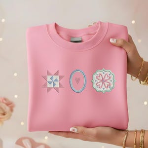 May include: A pink sweatshirt with three embroidered patches: a starburst, an oval with a heart, and a floral design. The patches are in shades of blue, pink, and green. The sweatshirt is held by a person.