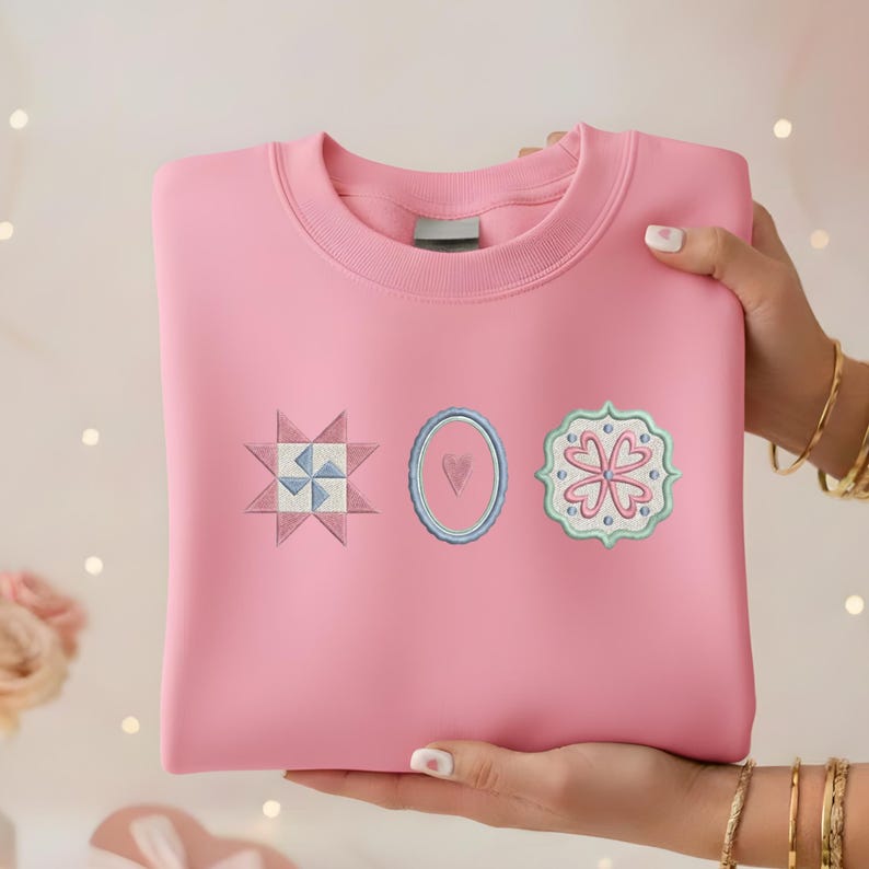 May include: A pink sweatshirt with three embroidered patches: a starburst, an oval with a heart, and a floral design. The patches are in shades of blue, pink, and green. The sweatshirt is held by a person.
