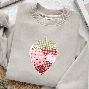 May include: A beige sweatshirt with a crew neck and a strawberry applique. The strawberry design is made of various fabric patterns, including gingham, polka dots, and floral prints in shades of red, pink, and green.