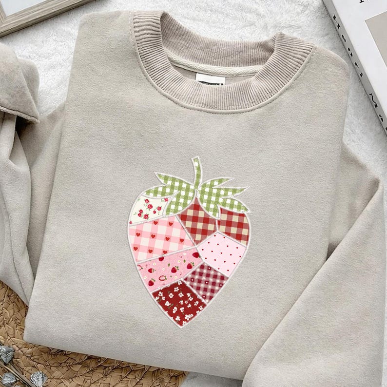 May include: A beige sweatshirt with a crew neck and a strawberry applique. The strawberry design is made of various fabric patterns, including gingham, polka dots, and floral prints in shades of red, pink, and green.
