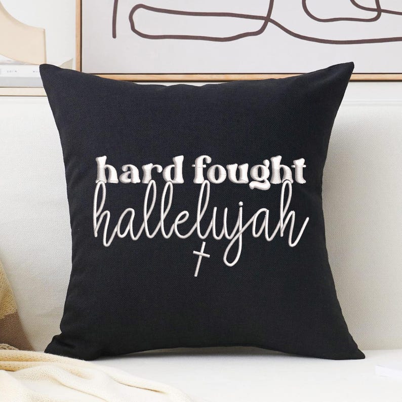 K&ouml;nnte beinhalten: Schwarzes quadratisches Kissen mit wei&szlig;em Text, der "hard fought hallelujah" in einer Schreibschriftart lautet. Unter dem Wort "hallelujah" befindet sich ein kleines Kreuz. Das Kissen liegt auf einem wei&szlig;en Sofa.