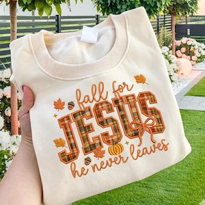 May include: A cream-coloured crewneck sweatshirt featuring the text "fall for JESUS he never leaves" in orange and brown plaid lettering. The design includes small orange pumpkin, acorn, and maple leaf illustrations with a bow accent.