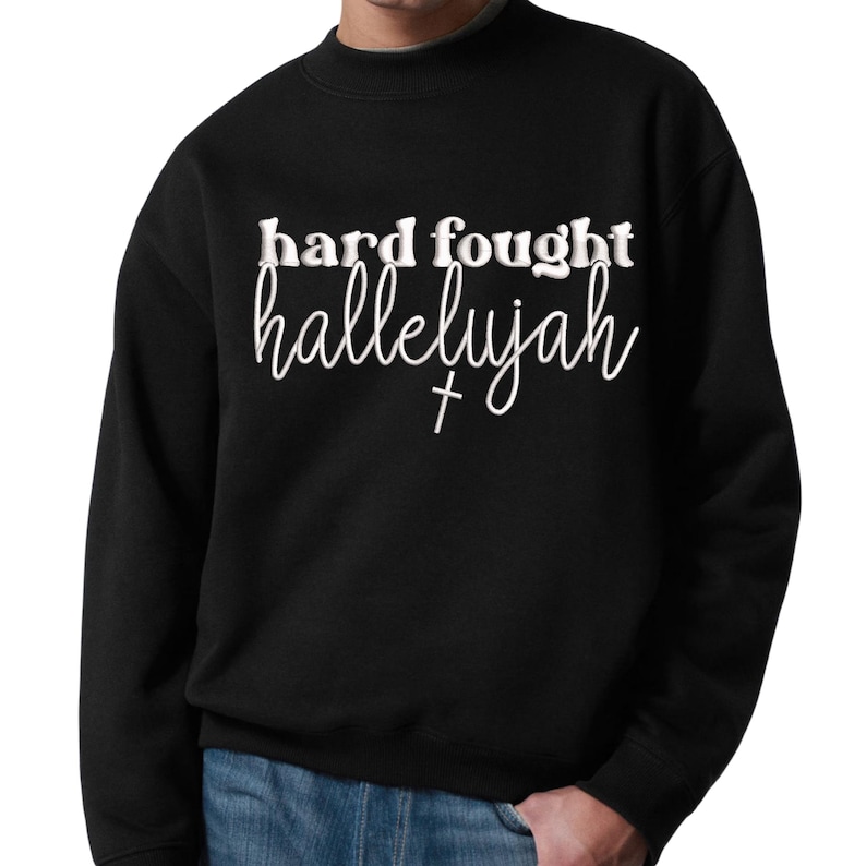 K&ouml;nnte beinhalten: Schwarzes Sweatshirt mit dem Schriftzug "hard fought hallelujah" in wei&szlig;er Schrift und Blockbuchstaben. Unter dem Wort "hallelujah" befindet sich ein kleines wei&szlig;es Kreuz. Das Sweatshirt hat einen Rundhalsausschnitt und lange &Auml;rmel.