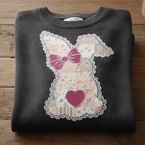 May include: A dark gray sweater with a decorative bunny applique. The bunny is made of patchwork fabric with floral patterns, a pink glitter bow, and a glitter heart. The applique is outlined with white lace.