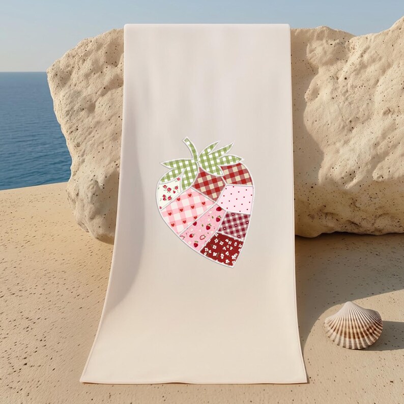 May include: A cream-colored beach towel featuring a large strawberry design. The strawberry is composed of various patchwork patterns in shades of red, pink, and green. A seashell rests nearby.