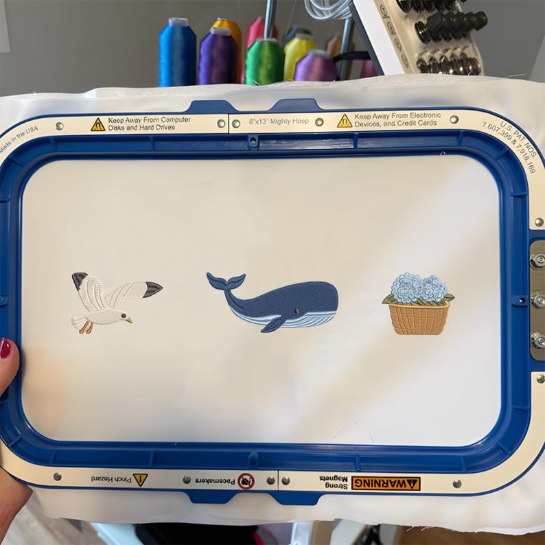 May include: A blue 8x13 inch Mighty Hoop embroidery frame holding white fabric with three embroidered designs: a white and gray seagull in flight, a blue whale, and a light blue hydrangea flower arrangement in a tan woven basket.