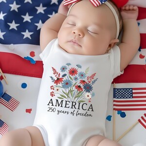 Puede incluir: Un body blanco para beb&eacute; con un dise&ntilde;o floral en rojo, blanco y azul, con la palabra "AMERICA" y "250 years of freedom" impresas debajo. El beb&eacute; est&aacute; acostado sobre una bandera estadounidense.