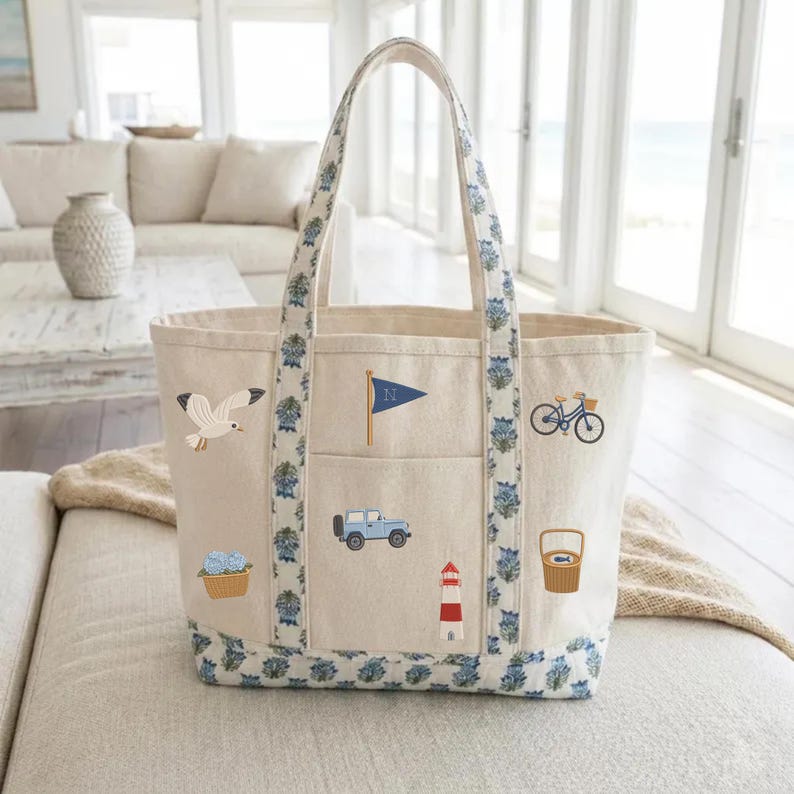 May include: A beige canvas tote bag with blue floral patterned trim and handles. The bag features embroidered icons including a seagull, a blue flag with the letter N, a bicycle, a blue SUV, a red and white lighthouse, and a basket of flowers.