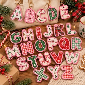 May include: A collection of twenty-six alphabet letter keychains, each featuring a pink border and various holiday-themed patterns including Christmas trees, red bows, gingerbread men, candy canes, and plaid prints, with metal rings.