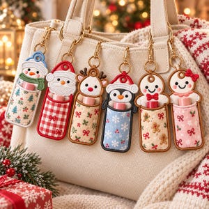 Op de afbeelding: Zes geborduurde stoffen lippenbalsemhouders met goudkleurige metalen clips bevestigd aan een crèmekleurige tas. De houders tonen figuren zoals een sneeuwman, de Kerstman, een rendier, een pinguïn, een peperkoekmannetje en een pop.