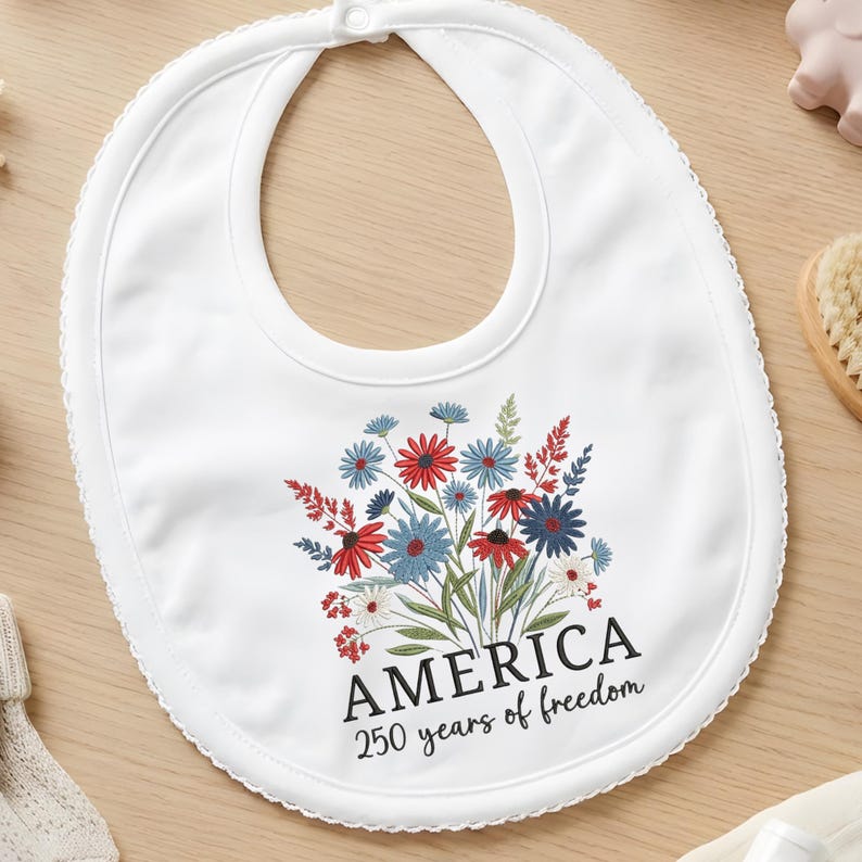 Puede incluir: Babero blanco para beb&eacute; con un dise&ntilde;o floral en rojo, azul y blanco. El babero presenta la palabra "AMERICA" y el texto "250 years of freedom" en negro. El babero tiene un borde festoneado y un cierre a presi&oacute;n.
