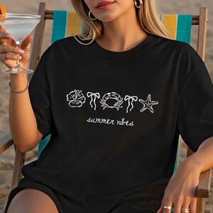 K&ouml;nnte beinhalten: Schwarzes T-Shirt mit wei&szlig;en Strichzeichnungen einer Blume, einer Schleife, einer Krabbe, einer Schleife und eines Seesterns. Die Worte "summer vibes" sind darunter in Schreibschrift geschrieben. Das T-Shirt wird von einer Person getragen, die in einem Strandstuhl sitzt.