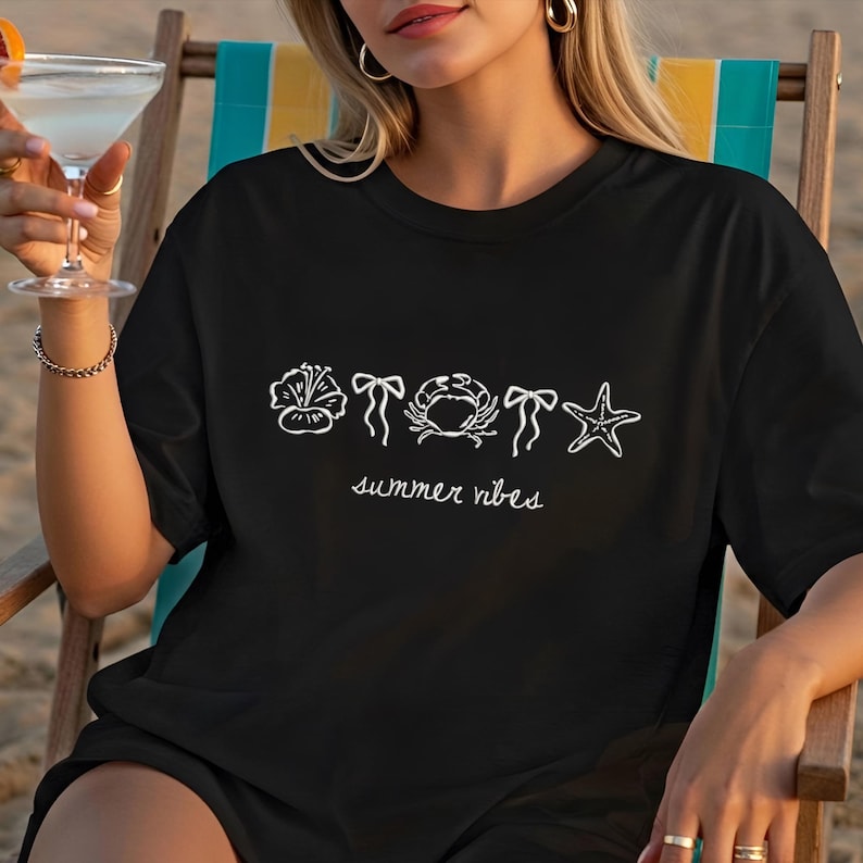 K&ouml;nnte beinhalten: Schwarzes T-Shirt mit wei&szlig;en Strichzeichnungen einer Blume, einer Schleife, einer Krabbe, einer Schleife und eines Seesterns. Die Worte "summer vibes" sind darunter in Schreibschrift geschrieben. Das T-Shirt wird von einer Person getragen, die in einem Strandstuhl sitzt.