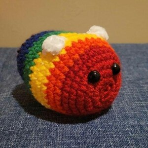 Könnte beinhalten: Ein handgehäkeltes, regenbogenfarbenes Bienenplüschtier mit schwarzen Perlenaugen und zwei kleinen weißen Pompon-Antennen. Die Biene ist rund und gestreift in Blau, Grün, Gelb, Orange und Rot. Ein weiches, kuscheliges Spielzeug.