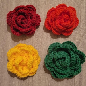May include: Four handmade crochet flowers in vibrant colors: red, orange, yellow, and green. Each flower is crafted with detailed petal layers, creating a realistic rose appearance. The flowers are arranged on a neutral gray surface.