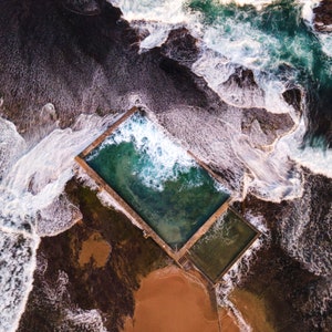 May include: An aerial view of a concrete swimming pool with turquoise water, surrounded by rocky terrain and crashing waves.