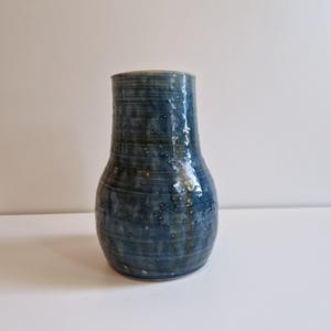 May include: A handcrafted, blue ceramic vase with a textured surface. The vase has a wide base that tapers to a narrower neck and a slightly flared rim. The color is a deep blue with subtle variations and speckles, giving it a unique, artisanal look.