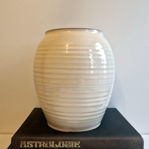 Könnte beinhalten: Weiße Keramikvase mit geripptem Design. Die Vase steht auf einem Stapel Bücher. Das oberste Buch hat den Schriftzug "ASTROLOGIE" auf dem Einband.