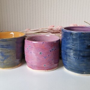 Può includere: Tre tazze in ceramica con diversi motivi di smalto. Le tazze sono in tonalità di blu, rosa e giallo. Le tazze sono disposte in fila su una superficie bianca.