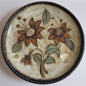 May include: A brown and white ceramic plate with a raised floral design. The plate has a brown rim and a label that reads "Gres Gregoire - Charente".