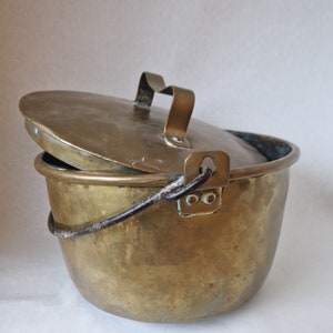 Puede incluir: Una olla de cobre antigua con tapa y asa de alambre. La olla tiene un aspecto desgastado y envejecido y es un gran ejemplo de utensilios de cocina antiguos.