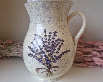 以下が含まれることがあります： ラベンダーの花柄が描かれた白い陶器のピッチャー。ピッチャーは斑点のある釉薬と取っ手があります。ラベンダーの花は紐でまとめられており、小さなタグには「J. Cagnon」と書いてあります。