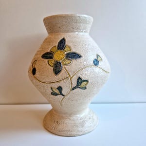 Puede incluir: Un jarrón de cerámica blanco con un diseño floral. El jarrón tiene una base ancha y un cuello estrecho. Las flores son azules, amarillas y verdes.