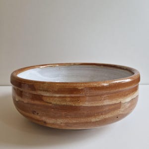 May include: A handmade ceramic bowl with a wide, shallow design. The exterior features horizontal bands of brown and tan hues, while the interior is a bright white. The bowl has a glossy finish, suggesting it's been glazed.
