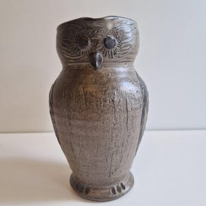 Puede incluir: Un jarrón artesanal de cerámica con forma de búho en tonos grises y marrones. El jarrón presenta detalles texturizados, incluyendo líneas talladas y rasgos faciales. El búho tiene ojos oscuros y circulares y un pequeño pico. La base tiene tres pequeños detalles en forma de garra.