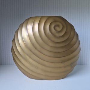Puede incluir: Un jarrón de metal dorado con un diseño en espiral. El jarrón tiene forma de concha.