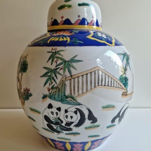 Puede incluir: Un jarrón de porcelana blanca con tapa, decorado con un diseño floral azul y amarillo. El jarrón presenta una escena de dos pandas en un bosque de bambú, con un puente y árboles en el fondo.