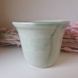 May include: A small, light green ceramic planter with a white glaze. The planter has a rounded bottom and a flared top.