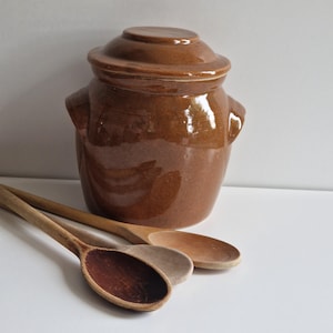 Puede incluir: Un tarro de cerámica marrón con tapa y dos cucharas de madera. El tarro tiene un acabado brillante y es de estilo tradicional.