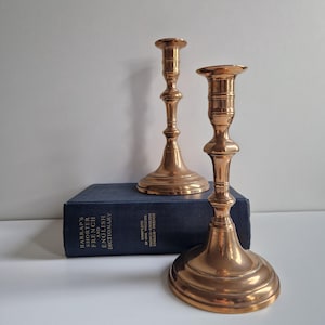 Op de afbeelding: Twee antieke messing kandelaars met een eenvoudig ontwerp. De kandelaars staan op een blauw boek met gouden letters.