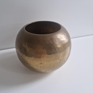 Puede incluir: Un macetero de latón martillado con forma redonda. El macetero tiene un acabado liso y brillante y una superficie ligeramente texturizada.